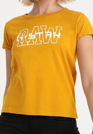 Vrouw die een mosterdgele T-shirt draagt met opgerolde mouwen en witte overlappende "RAW"-tekst op de borst, gecombineerd met zwarte broeken.