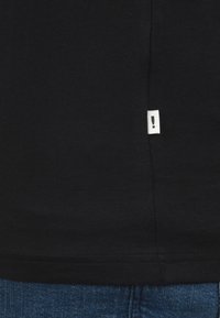 Camiseta negra de algodón con dobladillo recto y una pequeña etiqueta blanca con un signo de exclamación en el costado. Combinada con jeans de mezclilla azules.