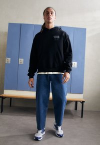 Sweatshirt à capuche noire avec poche avant, graphiques gris ; jean en denim bleu ample ; baskets blanches avec accents violets ; cadre dans une salle de sport.