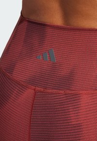 Røde mønstrede leggings med et struktureret design, der har en glat talje og et sort Adidas-logo øverst til venstre.