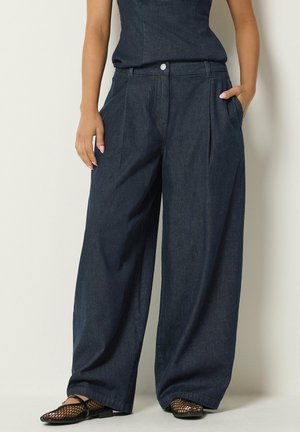 Persona con pantalones vaqueros oscuros de pierna ancha con bolsillos laterales y zapatillas negras de red, de pie contra una pared clara.