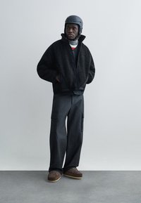 Veste en polaire noire zippée, associée à un pantalon cargo foncé. Un casque gris complète la tenue. Détails subtils sur des chaussures texturées marron.