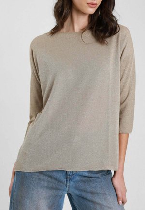 Donna che indossa un top beige scintillante a maniche lunghe tre quarti e jeans di denim azzurro, in piedi con le mani lungo i fianchi.