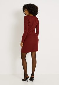 Robe ajustée bordeaux à manches longues, encolure ronde, et fermeture éclair au dos. Associée à des talons noirs et des collants transparents noirs.