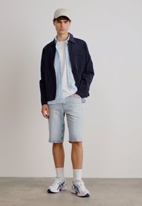 Giacca navy sopra una camicia azzurro chiaro, t-shirt bianca, pantaloni corti azzurri chiari e sneakers bianche con dettagli blu, il tutto su uno sfondo neutro.