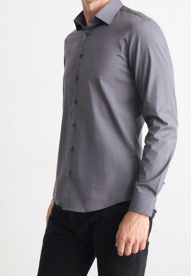 Homme portant une chemise grise ajustée à manches longues boutonnée, associée à un pantalon noir, sur un fond uni.