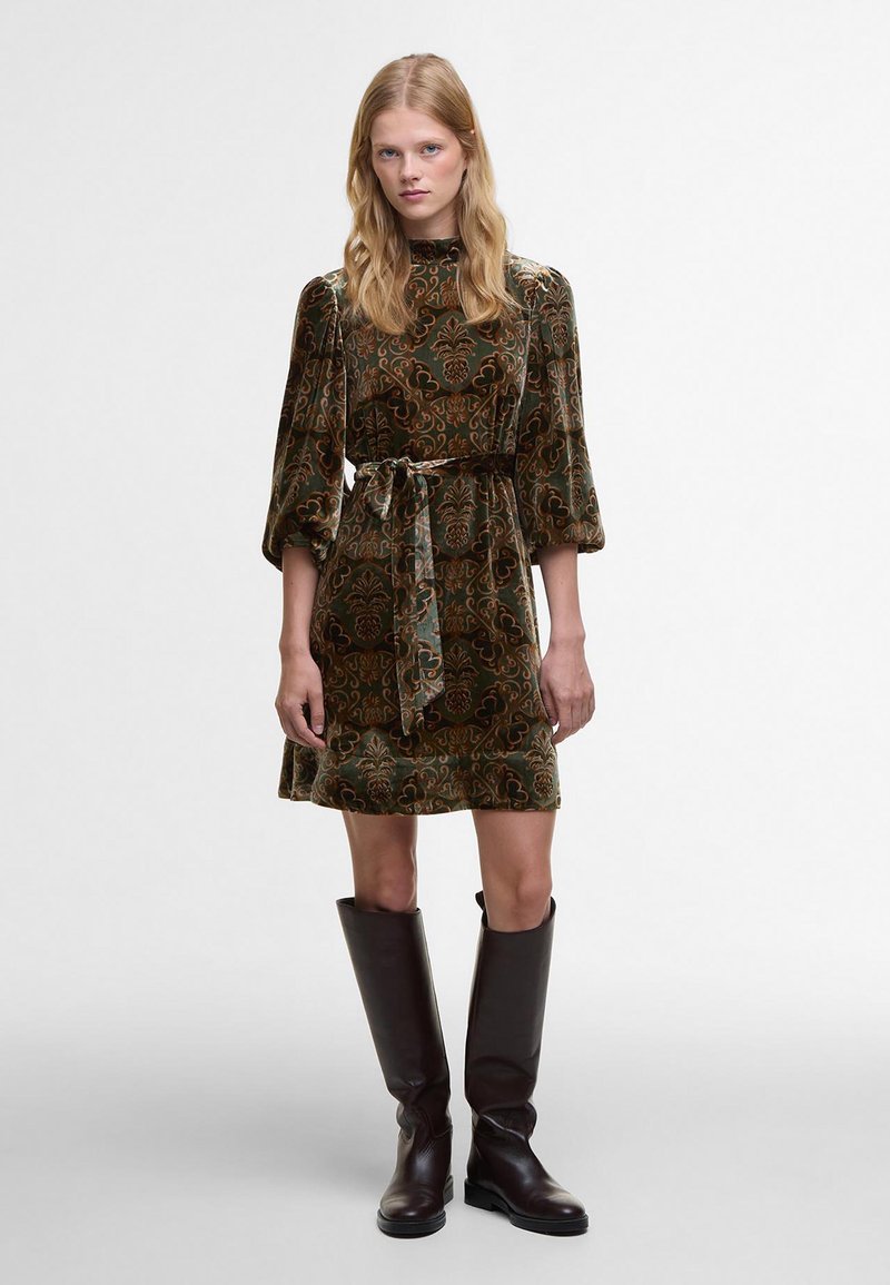 Βελούδινο πράσινο φόρεμα με λουλουδάτο μοτίβο, ψηλό λαιμόκοψι και φουσκωτά μανίκια, που είναι μαζεμένο στη μέση με ζώνη στο ίδιο χρώμα. Συνδυάζεται με καφέ μπότες μέχρι το γόνατο.