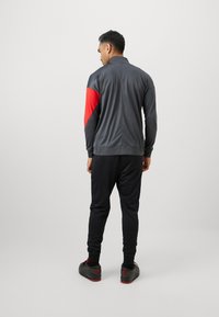 Grå træningsjakke med røde accenter, parret med sorte joggers. Jakken har en høj krave og er lavet af glat stof. Sorte sko fuldender looket.