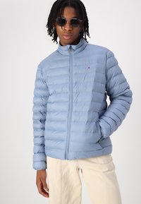 Lys blå puffjakke med horisontal quiltet design, glidelås i front, høy krage og sidelommer. Logo aksent på brystet.