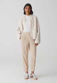 Romige fleecejack over een witte top, gecombineerd met beige corduroy broek en grijze schoenen met zwarte strepen. Soepele texturen en een relaxte pasvorm.
