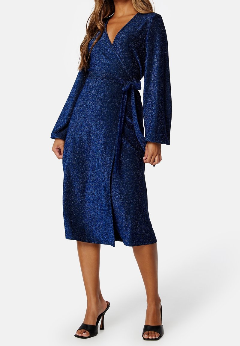 Femme portant une robe portefeuille bleu marine aux manches longues, longueur genoux, avec un lien sur le côté et des talons hauts noirs à bouts ouverts sur fond blanc.