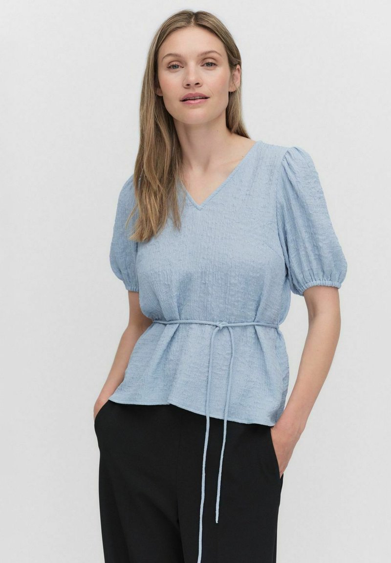 Ljusblå, rynkad tygblus med V-ringning och puffärmar, åtdragen i midjan med ett band. Matcher med svarta byxor.