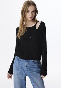 Pull en maille noire avec un léger col en V, comportant une fermeture à bouton unique. Assorti à un jean taille haute bleu clair.