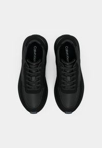 Μαύρα αθλητικά παπούτσια με λείο συνθετικό πάνω μέρος, υφή λεπτομέρειες, παχιές σόλες και κορδόνια, με λογότυπο ορατό στα εσωτερικά τους.