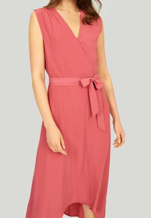 Bezruké korálové zavinovací šaty s výstřihem do V a zavázaným páskem v pase, které nosí žena na jednobarevném světle šedém pozadí.