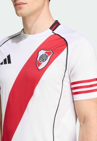 Hvid trøje med en rød diagonal stripe, sorte akcenter og ærmer med røde striber. Har et logo på brystet og en atletisk pasform.