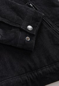 Gros plan sur la manche d'une veste en velours côtelé noir avec un bouton-pression argenté et une texture côtelée visible.
