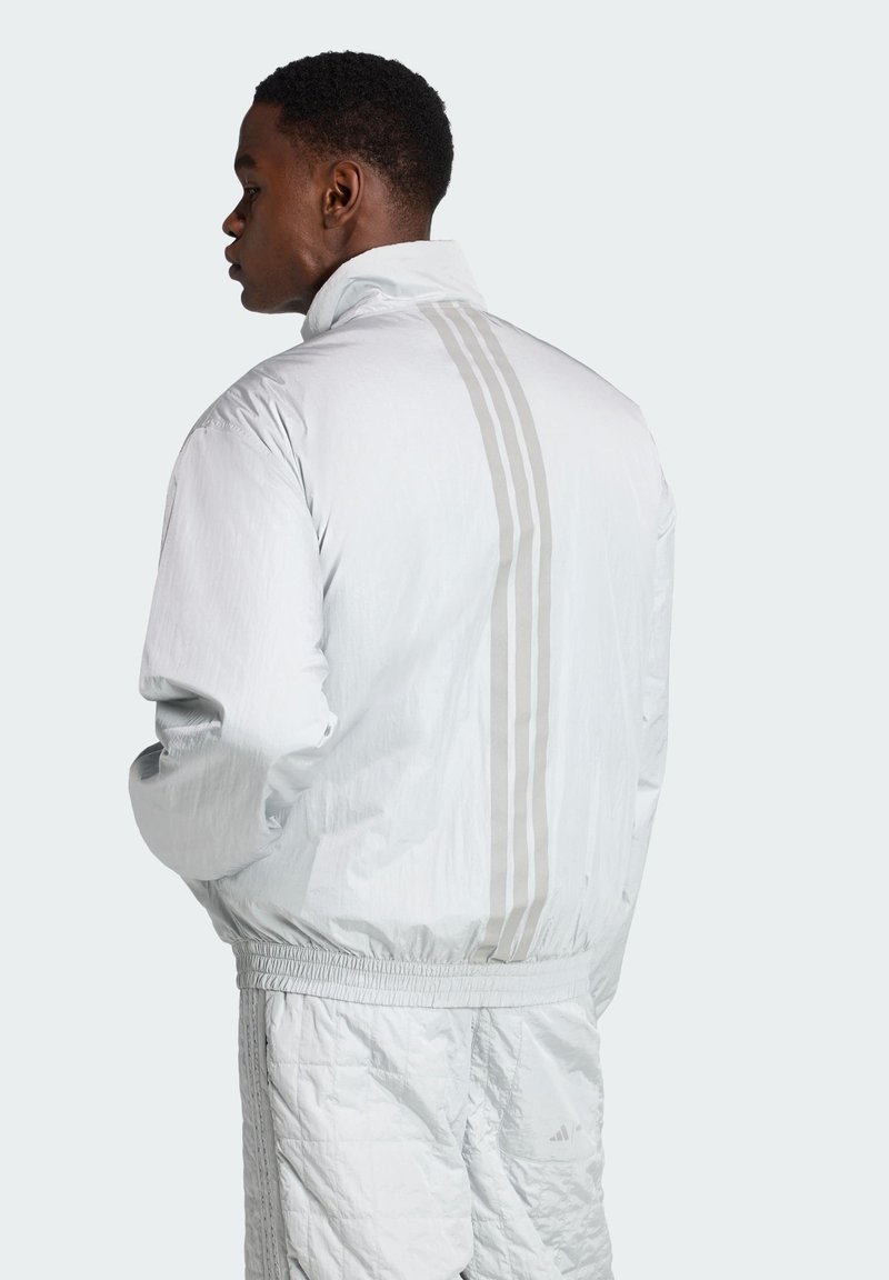 Chaqueta cortavientos gris claro con cuello alto, que presenta tres rayas verticales blancas en la parte posterior, hecha de un material liso y ligero.