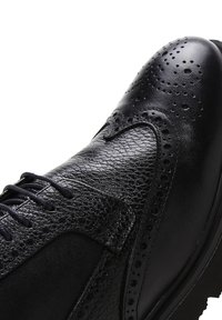 Chaussure en cuir noir présentant une combinaison de surfaces lisses et texturées, des perforations décoratives et un design à lacets.