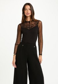 Top negro de manga larga con mangas transparentes de lunares y yugo de encaje. Combinado con pantalones negros de pierna ancha de talle alto, que presentan sutiles acentos de hardware.