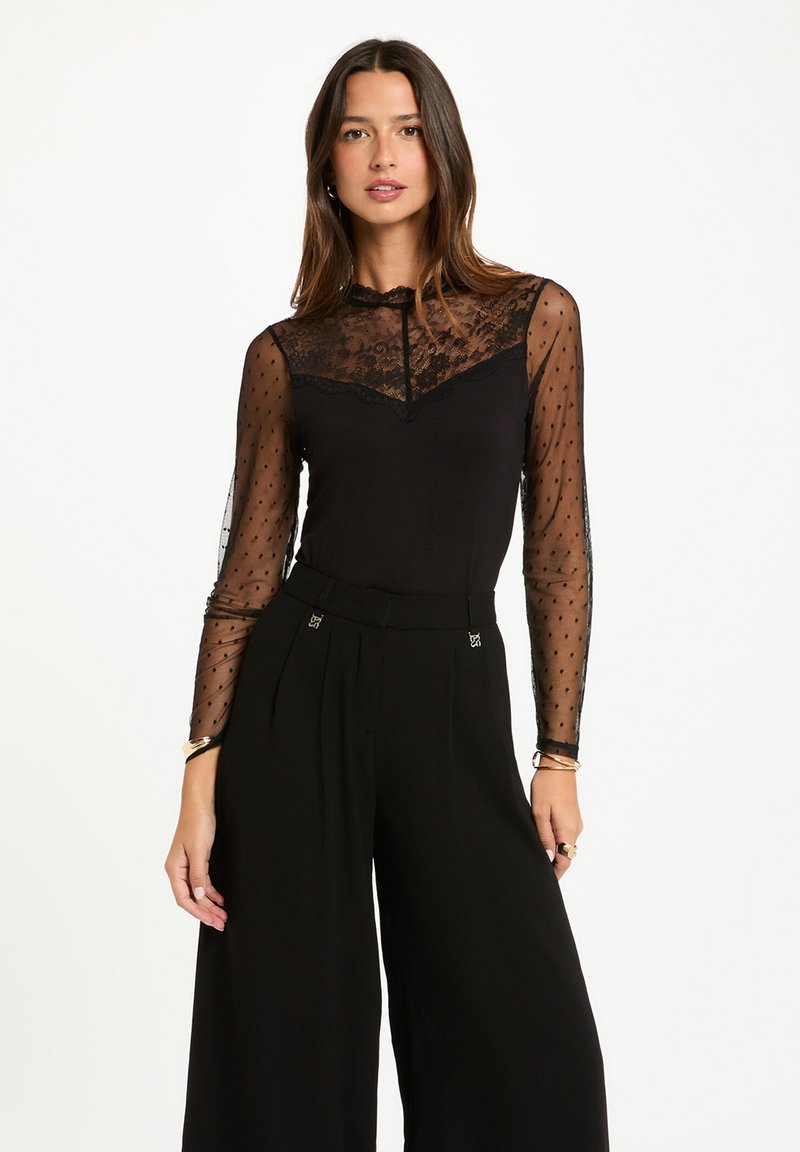 Top negro de manga larga con mangas transparentes de lunares y yugo de encaje. Combinado con pantalones negros de pierna ancha de talle alto, que presentan sutiles acentos de hardware.