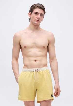 Ung mann som står mot en enkel bakgrunn, skjorteløs, iført lysegule badeshorts med snor og hendene i lommene.