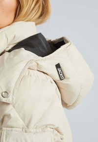Doudoune beige clair avec capuche, dotée d'une doublure noire, de poches zippées et d'une étiquette portant la mention "JACKIT" sur le col.