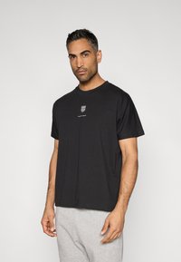 Schwarzes Baumwoll-T-Shirt mit kurzen Ärmeln, rundem Ausschnitt und einem kleinen weißen Logo auf der Brust. Der Stoff wirkt weich und glatt.