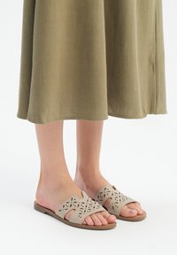 Beżowe klapki z wyciętymi wzorami, płaską podeszwą, otwartym noskiem i gładką fakturą, noszone z oliwkową spódnicą.