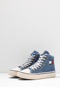 Vysoké tenisky vyrobené z modrého denimu se třepenými okraji, béžovými gumovými podrážkami, bílými špičkami a logem s červenými a modrými akcenty.