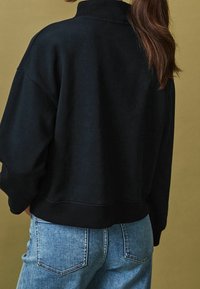 Woman with long brown hair wearing a black sweatshirt and blue denim jeans, standing against an olive green background.