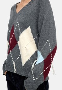 Szary sweter dzianinowy z dekoltem w serek, ozdobiony diamentowym wzorem w kolorze czerwonym, białym i jasnoniebieskim oraz frędzlami u dołu.
