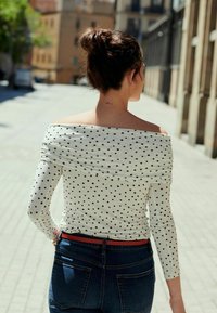 Balta off-the-shoulder palaidinė su juodais taškeliais, derinama su mėlynomis džinsinėmis kelnėmis ir raudonu diržu. Turi ilgus rankovėmis ir glotnią tekstūrą.