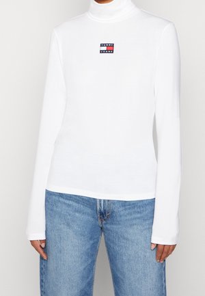 Osoba ubrana w biały, długi golf z małym logo Tommy Jeans na piersi oraz niebieskie dżinsy.