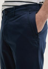 Pantalons en coton bleu marine à coupe slim, avec une poche avant et un détail zippé. Taille à bouton avec une couture de taille visible.