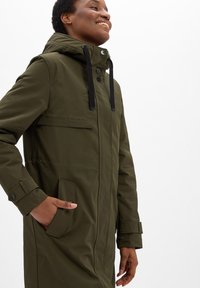 Person smiling, wearing an olive green hooded coat with black drawstrings, hand in pocket, standing against a plain white background.