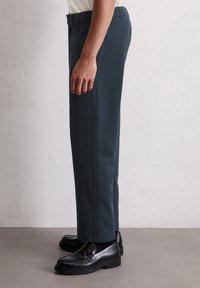 Pantalons ajustés teal en tissu texturé, présentant une couture latérale. Associés à des mocassins noirs, montrés de côté sur un fond clair.