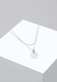 Collier en argent à double couche avec pendentif rond texturé sur une surface géométrique blanche sur fond gris.
