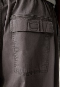 Poche en tissu gris foncé avec rabat et patch rectangulaire cousu sur un vêtement, montrant des coutures détaillées autour des bords.