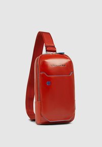 Bolso bandolera de cuero rojo con un acabado suave, que destaca por un detalle de cremallera azul y un logotipo. Correa ajustable para un uso cómodo.