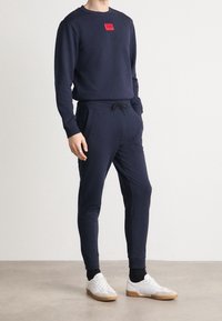 Navy sweatshirt met een rood logopatch, gecombineerd met bijpassende joggers met een elastische tailleband en trekkoord, aangevuld met witte sneakers.