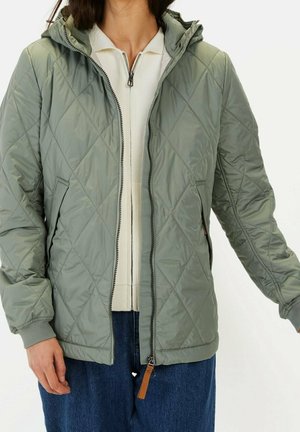 Personne portant une veste matelassée zippée vert clair par-dessus un haut blanc et un jean bleu, debout, les bras légèrement écartés.