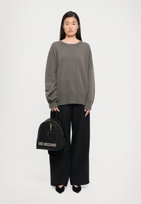 Woman wearing oversized gray sweater and wide-leg black pants, holding black Love Moschino backpack with gold zipper and logo.