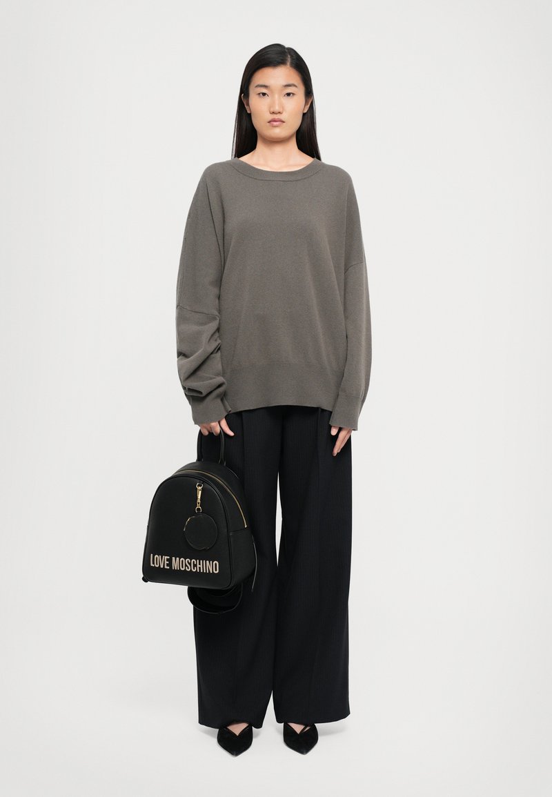 Woman wearing oversized gray sweater and wide-leg black pants, holding black Love Moschino backpack with gold zipper and logo.