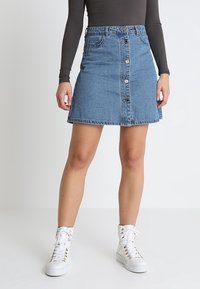 Jeans A-Linien-Rock, hellblau, mit Knopfverschluss, fünf Taschen und Kontrastnähten. Kombiniert mit weißen High-Top-Sneakern.