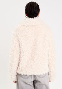 Veste en fausse fourrure douce et légère de couleur rose pâle, avec un col, des manches longues et un ourlet court. Matériau texturé et doux. Portée avec un jean gris clair.