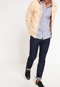 Beige lightweight jacket, blue and white striped button-up shirt, dark denim jeans, and black slip-on shoes with white soles.