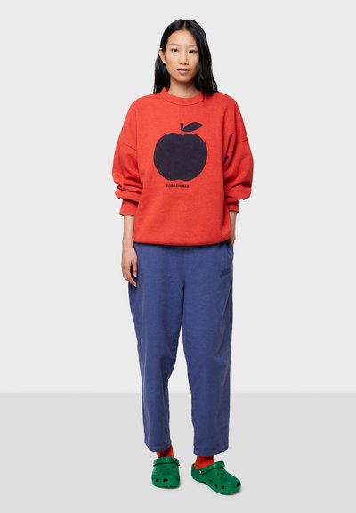 Rød sweatshirt med et æblegrafik, parret med blå sweatpants. Grønlige klip-klapper iført med røde sokker. Casual, afslappet pasform, blødt stof.