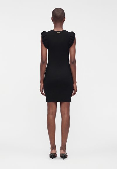 Femme portant une robe noire ajustée avec des manches courtes à volants, vue de dos, debout devant un fond blanc uni.