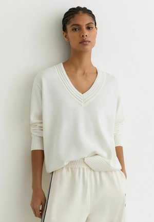 Vrouw met gevlochten haar, gekleed in een witte V-hals trui en witte broek met zwarte zijstrepen, staand tegen een witte muur.